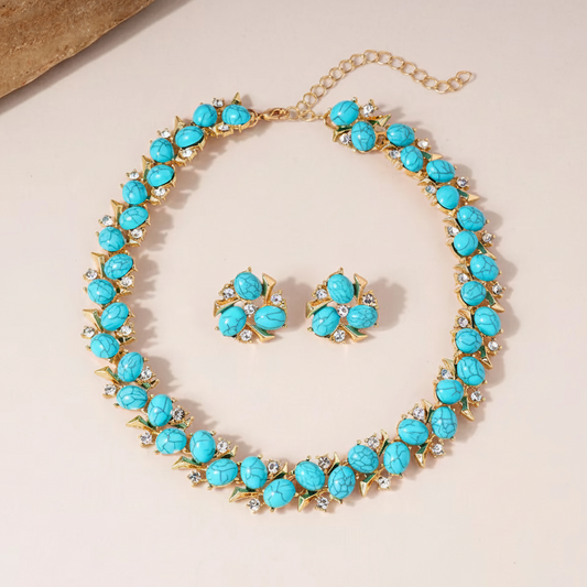 A gold-tone statement necklace and matching stud earrings set with faceted turquoise-blue stone centers and small clear rhinestone accents, arranged on a light beige background.