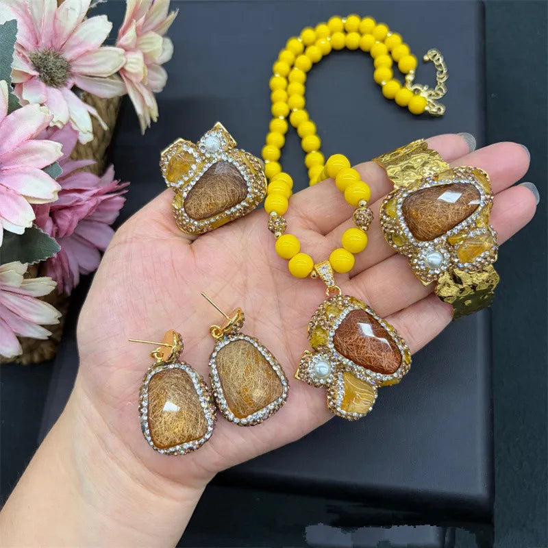 A gold jewelry set with a beaded yellow necklace, bracelet, earrings, and pendant, each featuring a large brown and gold textured stone surrounded by clear rhinestones, held in a hand against a black background with flowers.