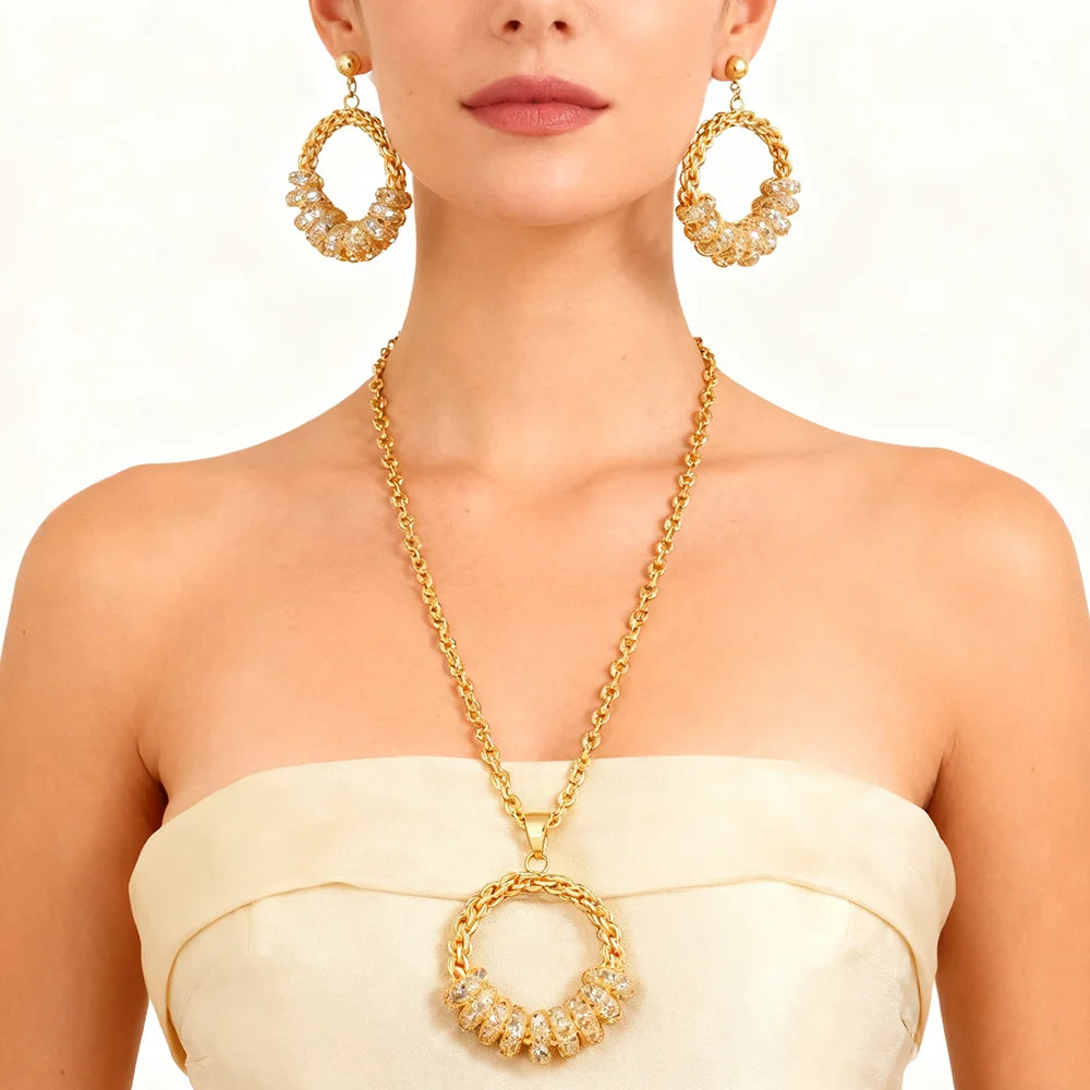 A woman wearing a gold necklace with a large circular pendant and matching drop earrings, styled with a cream strapless top against a plain light background.