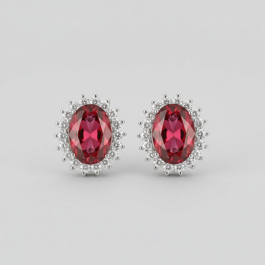A pair of oval red gemstone stud earrings with silver-toned halo settings, displayed on a pink velvet jewelry box.