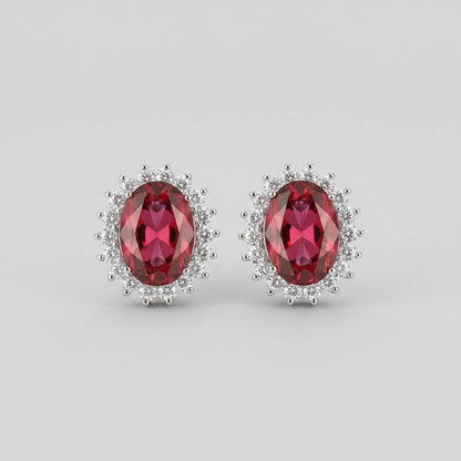 A pair of oval red gemstone stud earrings with silver-toned halo settings, displayed on a pink velvet jewelry box.
