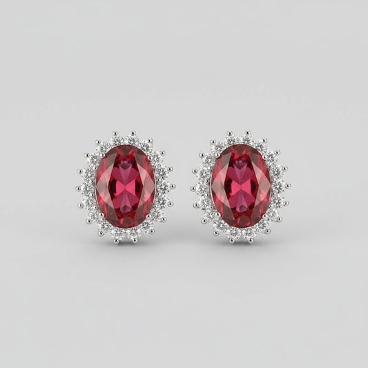 A pair of oval red gemstone stud earrings with silver-toned halo settings, displayed on a pink velvet jewelry box.