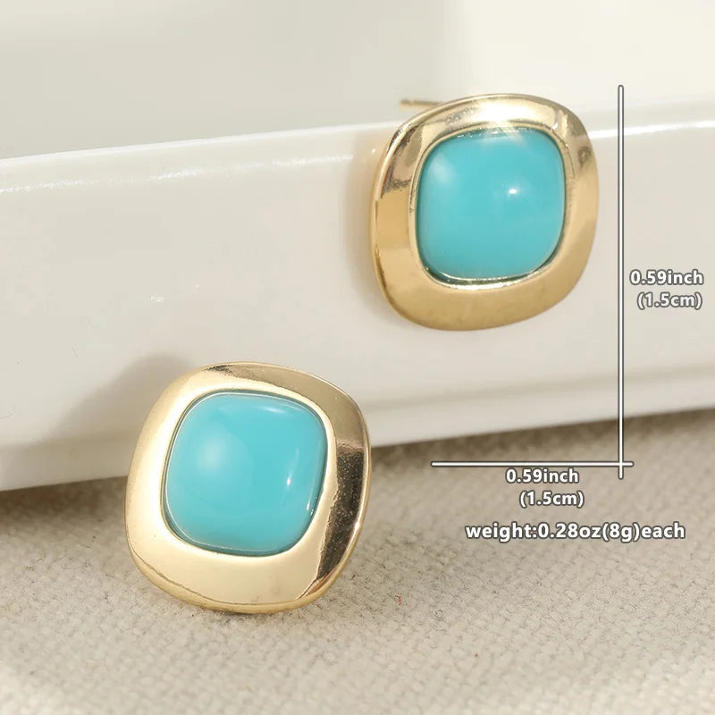 A pair of gold square stud earrings with rounded turquoise blue centers, displayed on a white tray and textured beige fabric. Measurement lines and text indicate dimensions of 0.