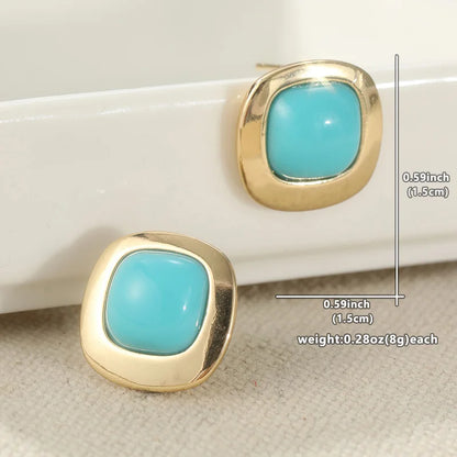 A pair of gold square stud earrings with rounded turquoise blue centers, displayed on a white tray and textured beige fabric. Measurement lines and text indicate dimensions of 0.