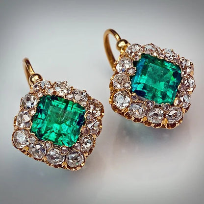 A pair of gold hoop earrings with large square-cut emerald-cut green gemstones surrounded by a halo of clear round-cut stones, shown on a neutral light gray background.