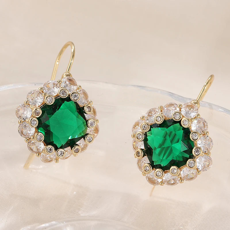 A pair of gold-tone drop earrings with a large emerald-cut green center stone surrounded by a halo of clear rhinestones, displayed on a clear glass surface against a warm beige background.