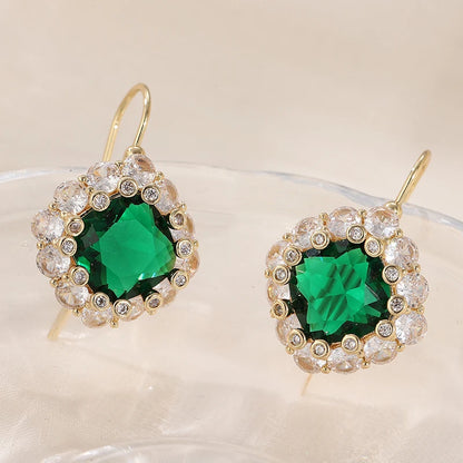 A pair of gold-tone drop earrings with a large emerald-cut green center stone surrounded by a halo of clear rhinestones, displayed on a clear glass surface against a warm beige background.