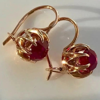 A pair of rose gold drop earrings with round deep red gemstones in a claw setting, displayed on a light off-white background.