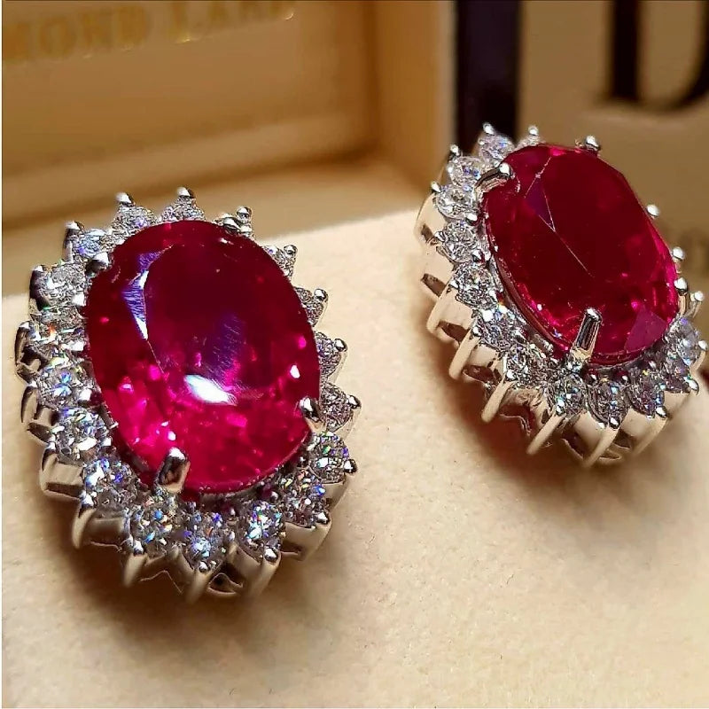 A pair of oval-cut red gemstone stud earrings with silver-toned halo settings and small clear accent stones, displayed on a beige surface with a blurred jewelry box in the background.