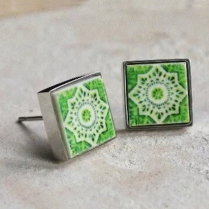 A pair of square stud earrings with green and white mandala-style inlay designs set in silver-tone metal, resting on a light textured surface.