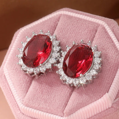 A pair of oval red gemstone stud earrings with silver-toned halo settings, displayed on a pink velvet jewelry box.