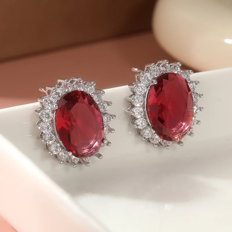 A pair of oval stud earrings with large deep red center stones surrounded by small clear stones in silver-toned settings, displayed on a white surface.
