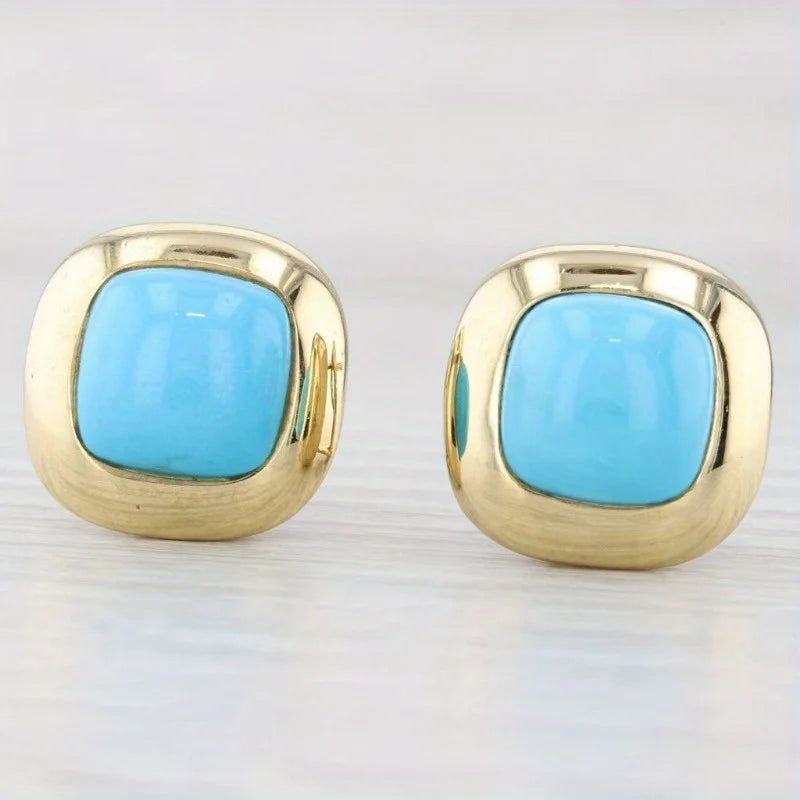A pair of gold square stud earrings with rounded corners and a central light blue cabochon stone, displayed on a light gray surface.