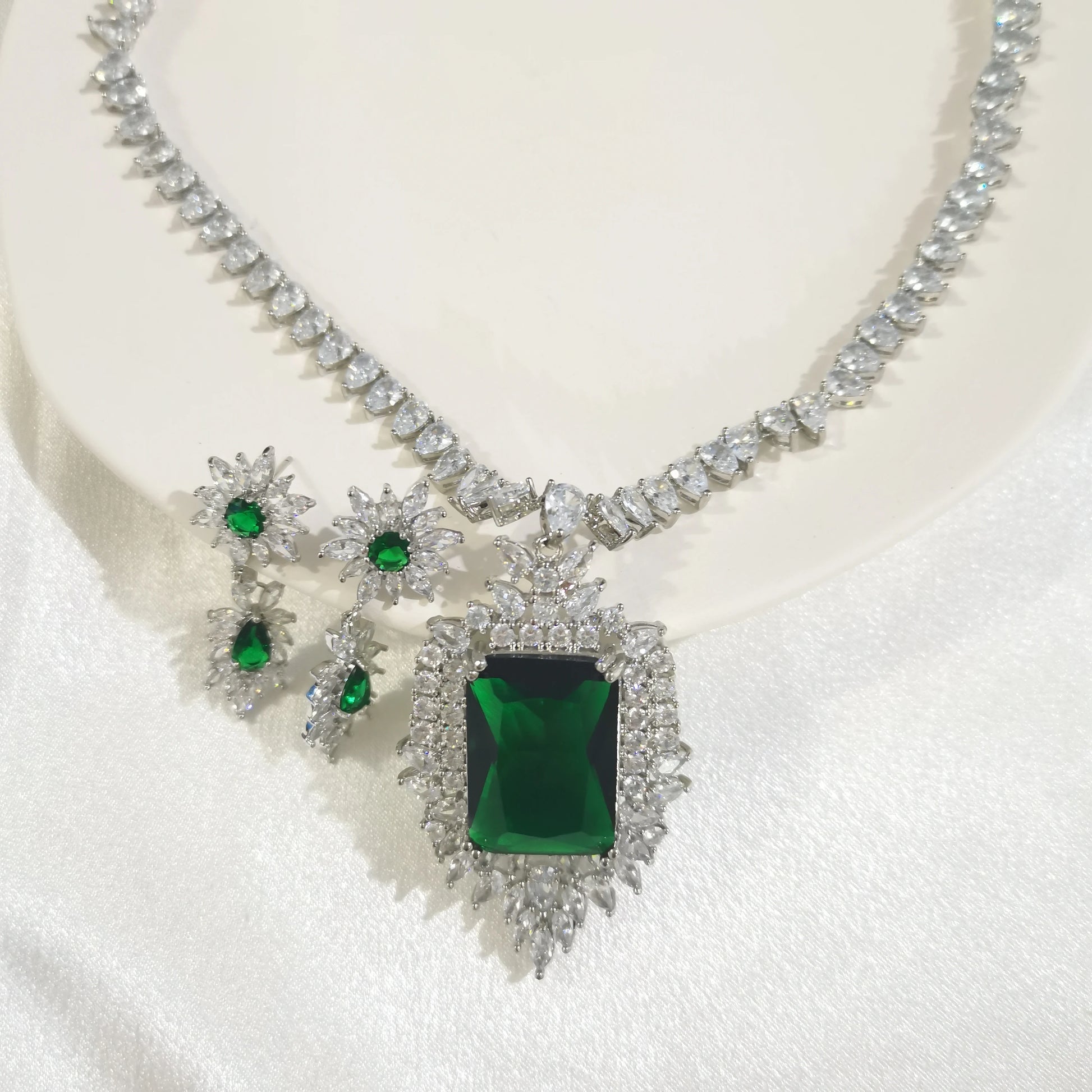 A silver-toned jewelry set featuring a necklace and matching earrings, decorated with clear crystals and green gemstones, displayed on a white satin surface.