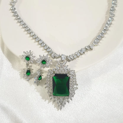 A silver-toned jewelry set featuring a necklace and matching earrings, decorated with clear crystals and green gemstones, displayed on a white satin surface.
