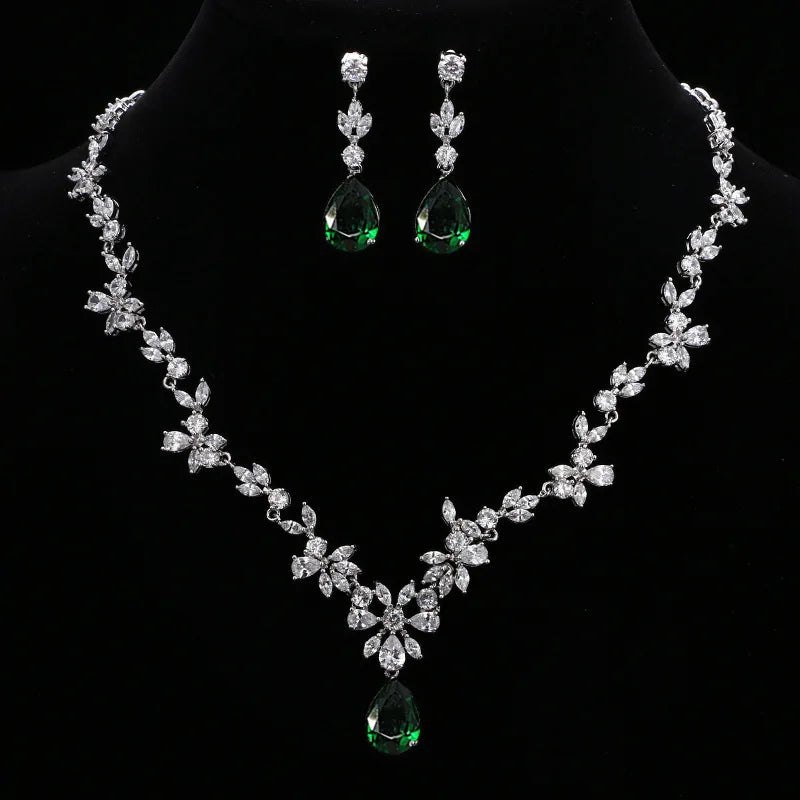 A silver-tone floral necklace and matching drop earrings displayed on a black jewelry bust. The necklace and earrings are set with clear stones and feature green teardrop gemstones at the center and bottom.