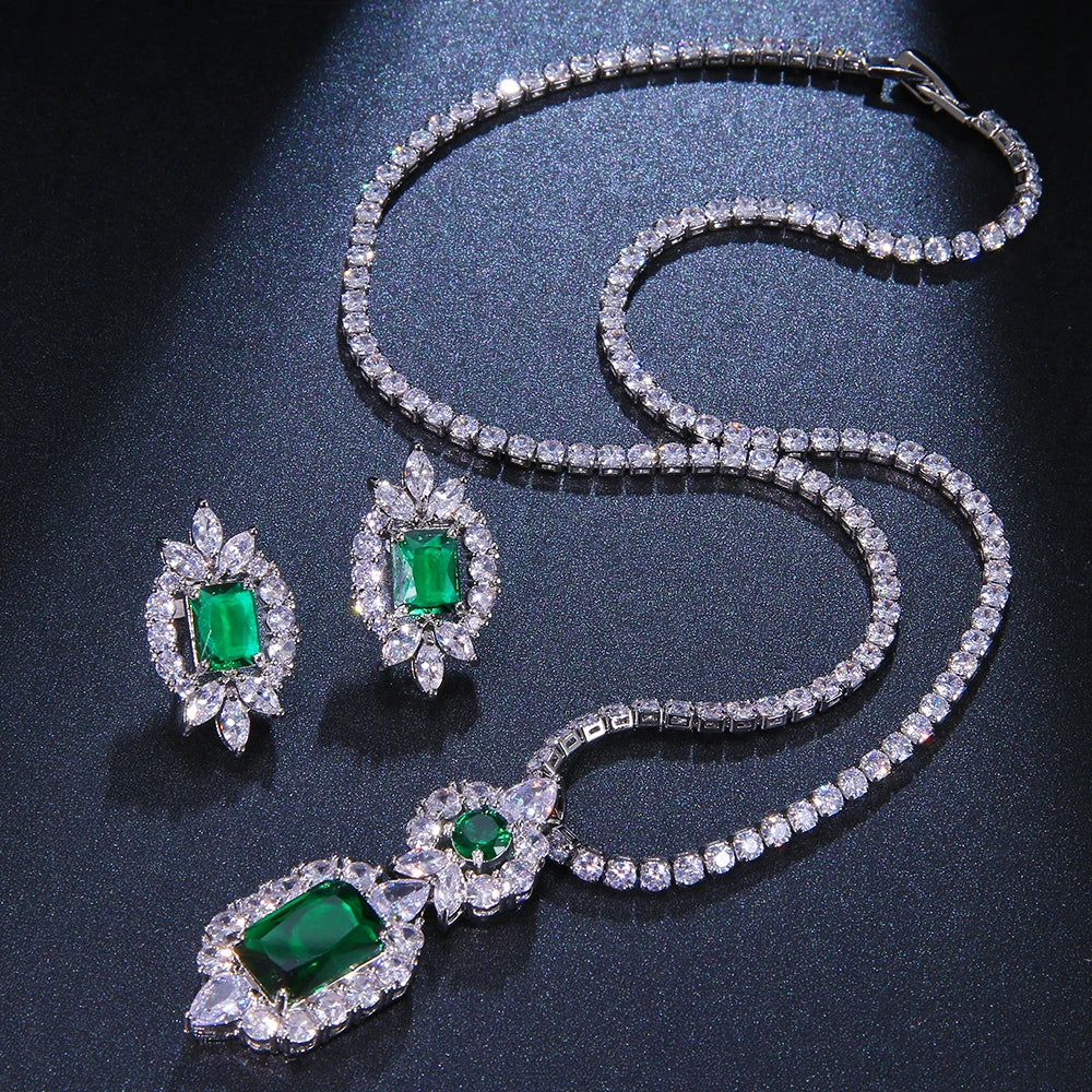 A silver-tone jewelry set featuring a necklace and matching earrings, both adorned with clear crystals and vibrant green emerald-cut gemstones, displayed on a dark textured background.
