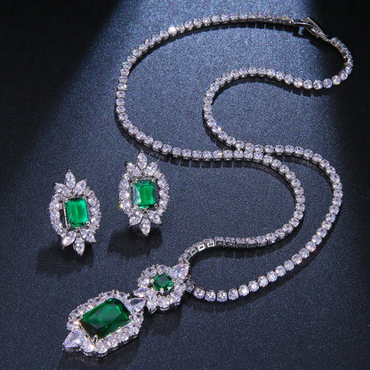 A silver-tone jewelry set featuring a necklace and matching earrings, both adorned with clear crystals and vibrant green emerald-cut gemstones, displayed on a dark textured background.