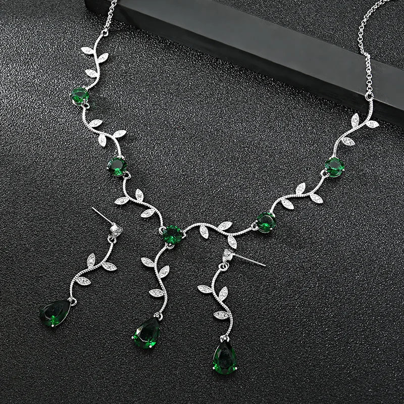Silver vine necklace and matching drop earrings with green teardrop gemstones and small clear stones, displayed on a dark textured surface.