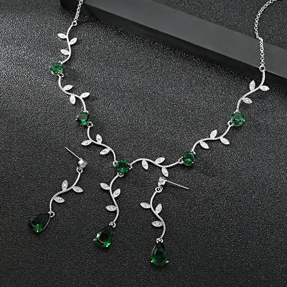 Silver vine necklace and matching drop earrings with green teardrop gemstones and small clear stones, displayed on a dark textured surface.