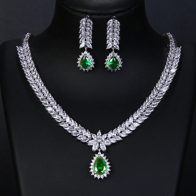 A silver-tone jewelry set featuring a leaf-patterned necklace and matching drop earrings, both adorned with clear crystals and a vibrant green teardrop gemstone at the center, displayed on a black mannequin bust.