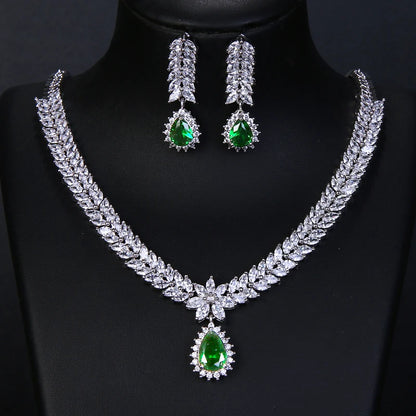 A silver-tone jewelry set featuring a leaf-patterned necklace and matching drop earrings, both adorned with clear crystals and a vibrant green teardrop gemstone at the center, displayed on a black mannequin bust.