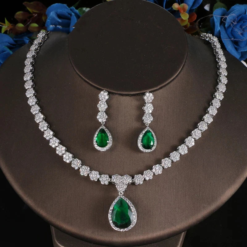 A jewelry set featuring a silver-tone necklace and matching drop earrings with emerald-cut green gemstones and clear accent stones, displayed on a dark brown jewelry bust.