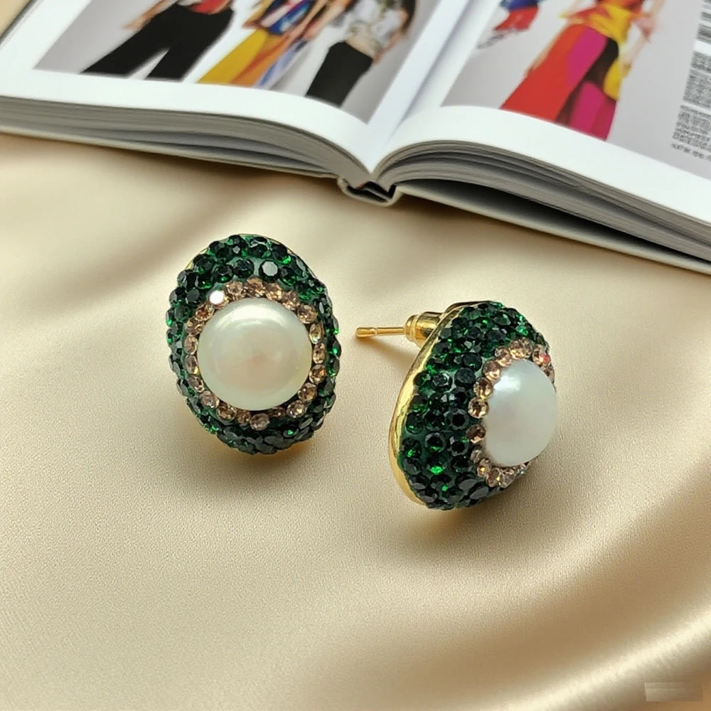 Pair of earrings with green gemstones and a pearl on a beige surface, with an open magazine in the background.