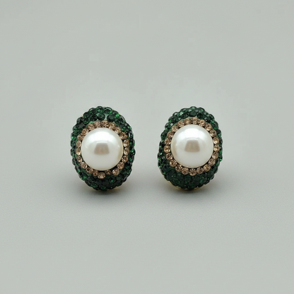 A pair of oval stud earrings featuring a central white pearl surrounded by a halo of dark green stones and a border of small clear crystals, set against a plain light gray background.