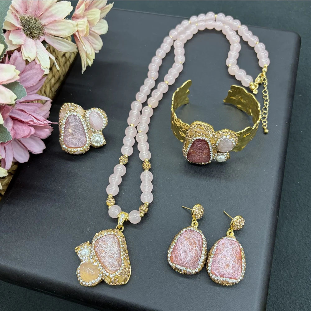 A jewelry set displayed on a dark gray surface, including a double-strand pink bead necklace with a large pink stone pendant, a gold cuff bracelet with a pink stone, matching drop earrings, and a small matching stud earring. Pink flowers are arranged on the left side of the frame.