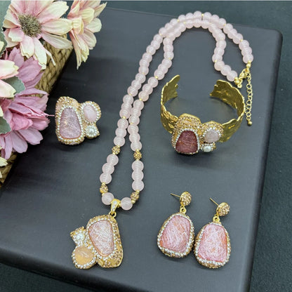 A jewelry set displayed on a dark gray surface, including a double-strand pink bead necklace with a large pink stone pendant, a gold cuff bracelet with a pink stone, matching drop earrings, and a small matching stud earring. Pink flowers are arranged on the left side of the frame.