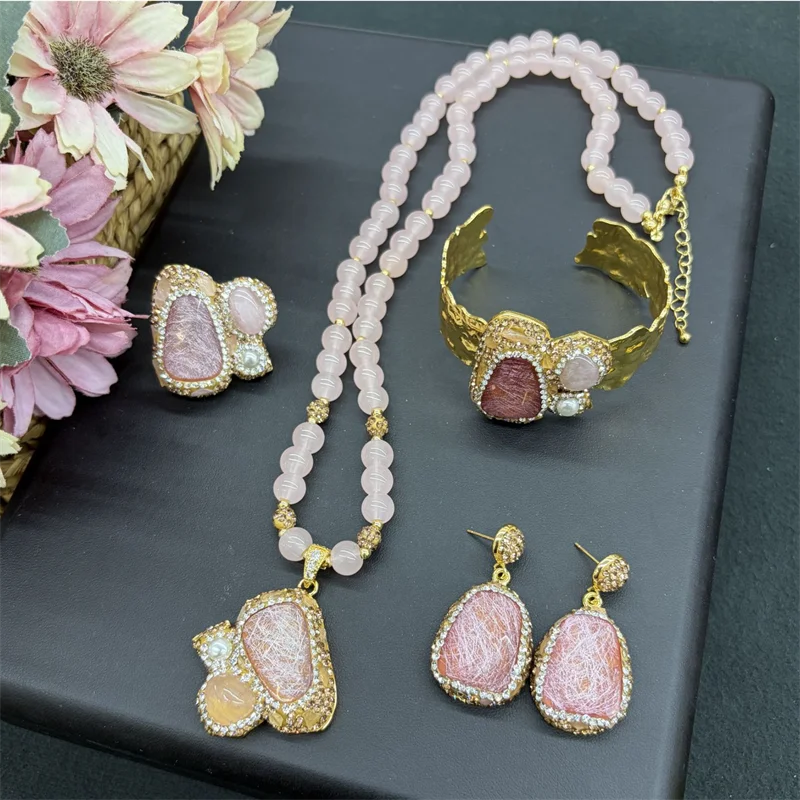 A jewelry set displayed on a dark gray surface, featuring a pink stone necklace, matching earrings, and a gold cuff bracelet. The pink stones are set in gold with clear rhinestone-like accents.