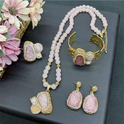 A jewelry set displayed on a dark gray surface, featuring a pink stone necklace, matching earrings, and a gold cuff bracelet. The pink stones are set in gold with clear rhinestone-like accents.