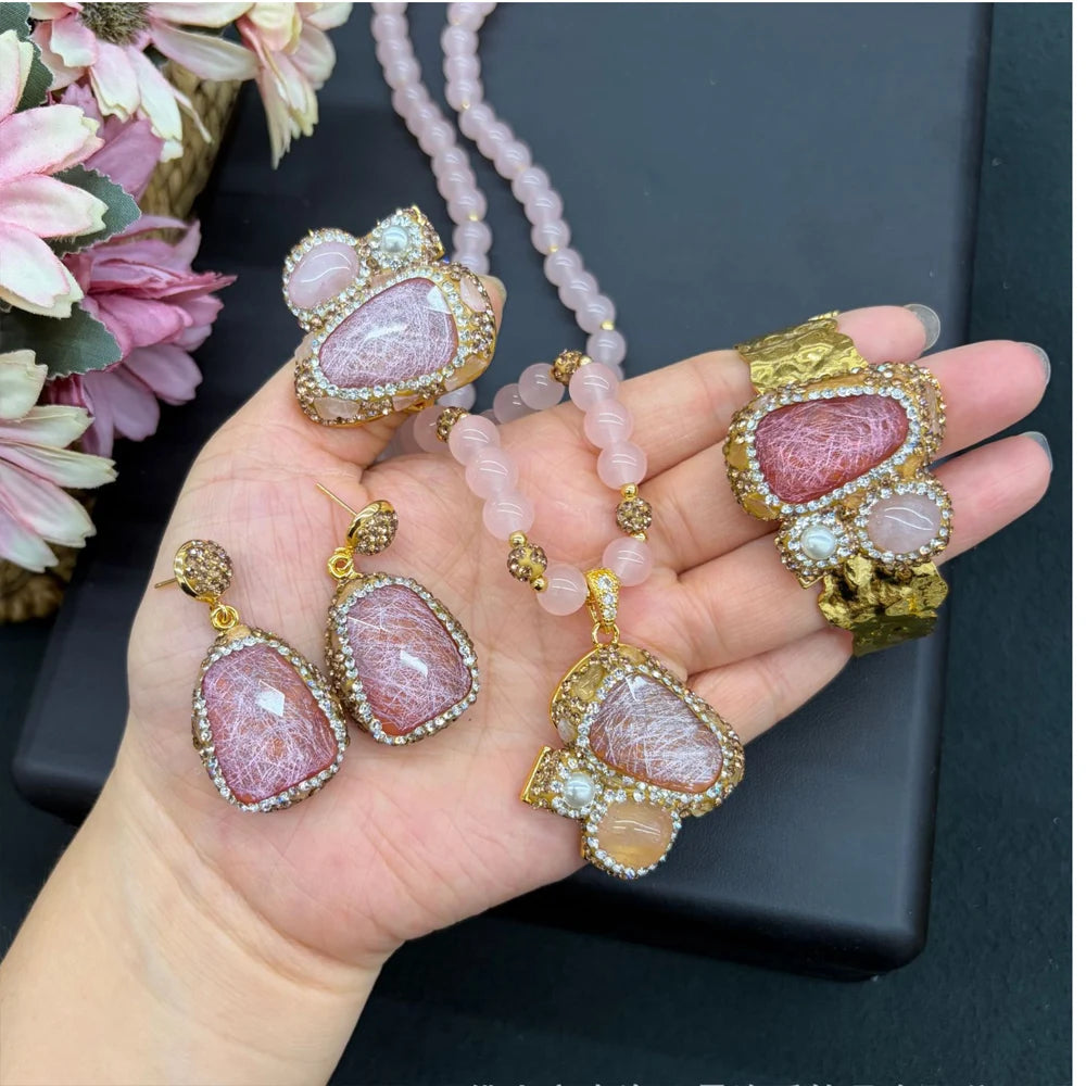 A hand holding a jewelry set featuring a pink stone pendant necklace, matching drop earrings, and a wide bracelet, all with gold-toned settings and pink beads.