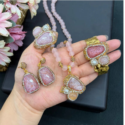 A hand holding a jewelry set featuring a pink stone pendant necklace, matching drop earrings, and a wide bracelet, all with gold-toned settings and pink beads.