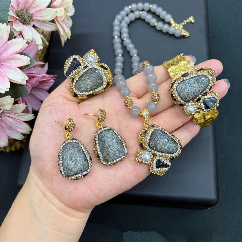 A jewelry set displayed on a hand, featuring a beaded necklace, a large pendant, a matching ring, and two earrings. The jewelry has gray stone-like centers with golden borders, clear rhinestones, and small black stone accents.