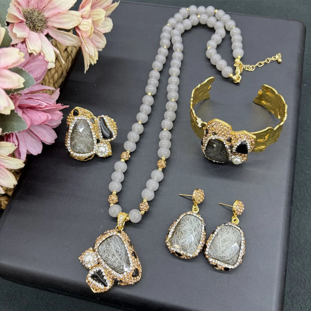 A jewelry set displayed on a dark gray box, featuring a beaded necklace, matching cuff bracelet, earrings, and a statement piece. The jewelry has gold-tone metal settings with gray and black textured stones, accented with small clear rhinestones.