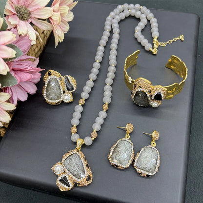 A jewelry set displayed on a dark gray box, featuring a beaded necklace, matching cuff bracelet, earrings, and a statement piece. The jewelry has gold-tone metal settings with gray and black textured stones, accented with small clear rhinestones.