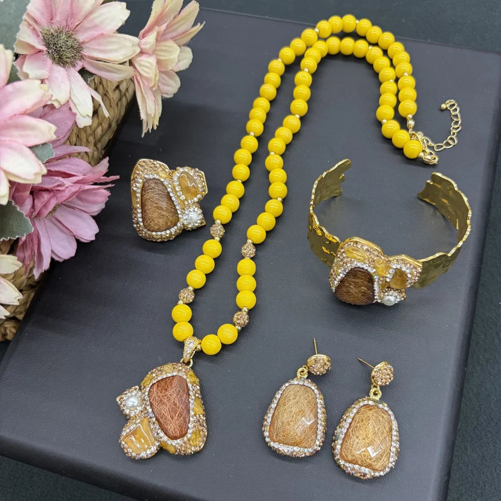A gold jewelry set featuring a beaded necklace, cuff bracelet, earrings, and ring, each decorated with a central brown stone surrounded by clear rhinestones, displayed on a dark gray surface with pink flowers in the corner.