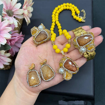 A gold jewelry set with a beaded yellow necklace, bracelet, earrings, and pendant, each featuring a large brown and gold textured stone surrounded by clear rhinestones, held in a hand against a black background with flowers.