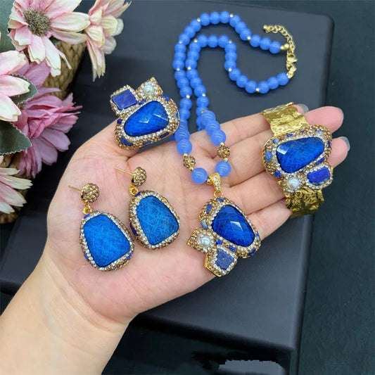 A jewelry set displayed on a person's hand, featuring a blue beaded necklace, a gold-tone ring, and matching drop earrings with large blue stones and gold borders.