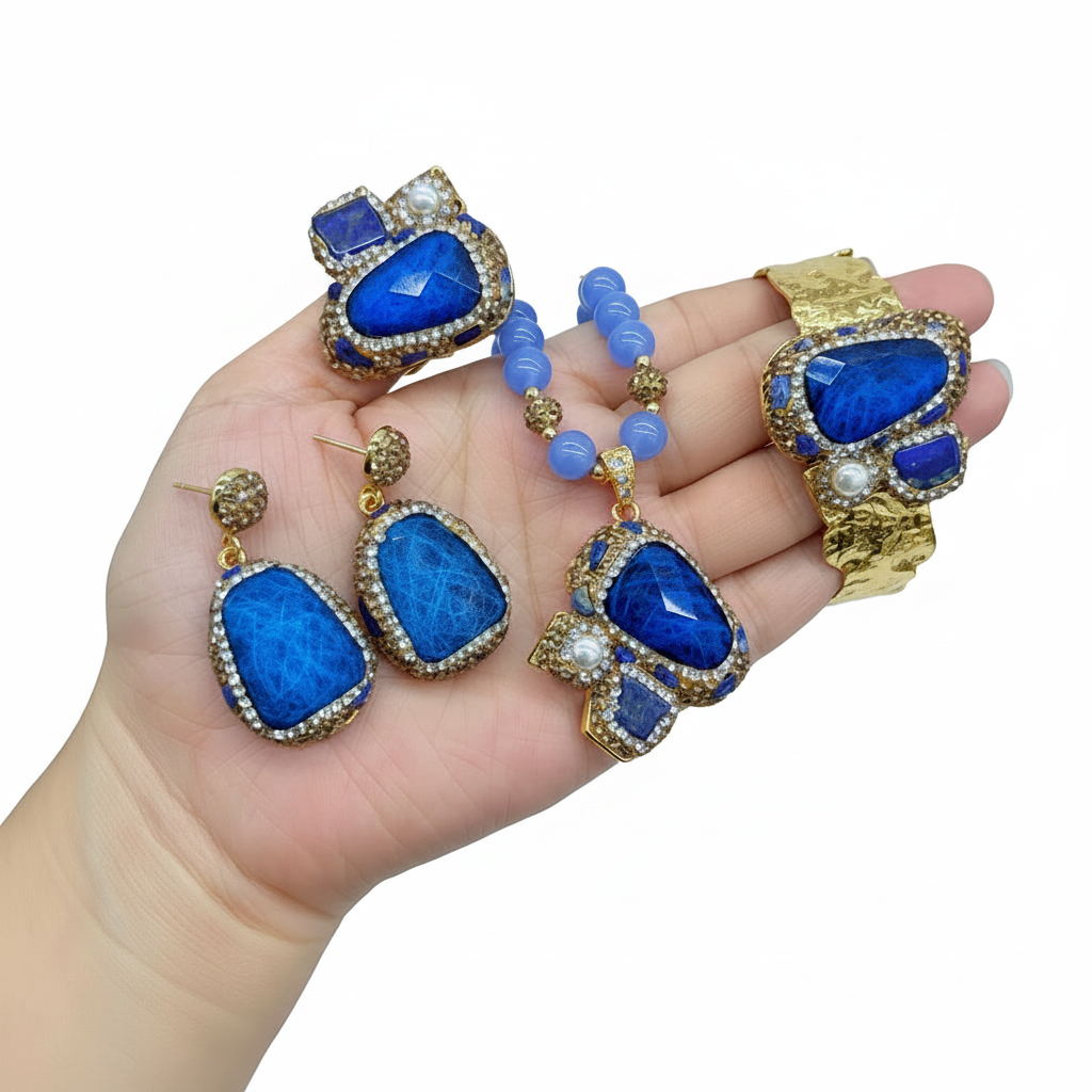 A jewelry set displayed on a hand, featuring a gold-tone bracelet, matching earrings, a beaded necklace, and decorative charms, all embellished with vibrant blue stones and small white pearl-like accents.