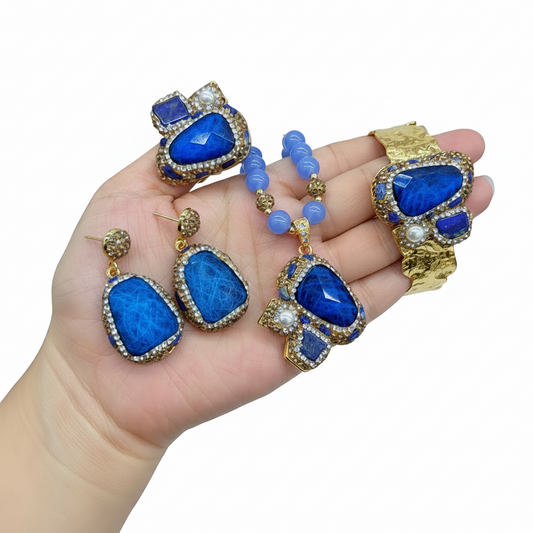 A jewelry set displayed on a hand, featuring a gold-tone bracelet, matching earrings, a beaded necklace, and decorative charms, all embellished with vibrant blue stones and small white pearl-like accents.