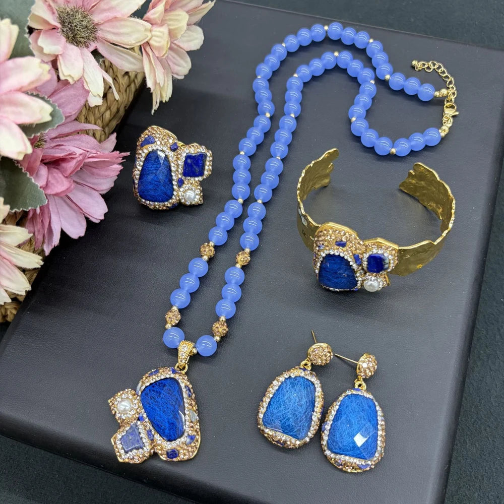 A gold-plated jewelry set featuring a beaded necklace, matching earrings, a cuff bracelet, and a ring, all decorated with large blue stones and small clear rhinestones, displayed on a dark gray surface next to pink flowers.