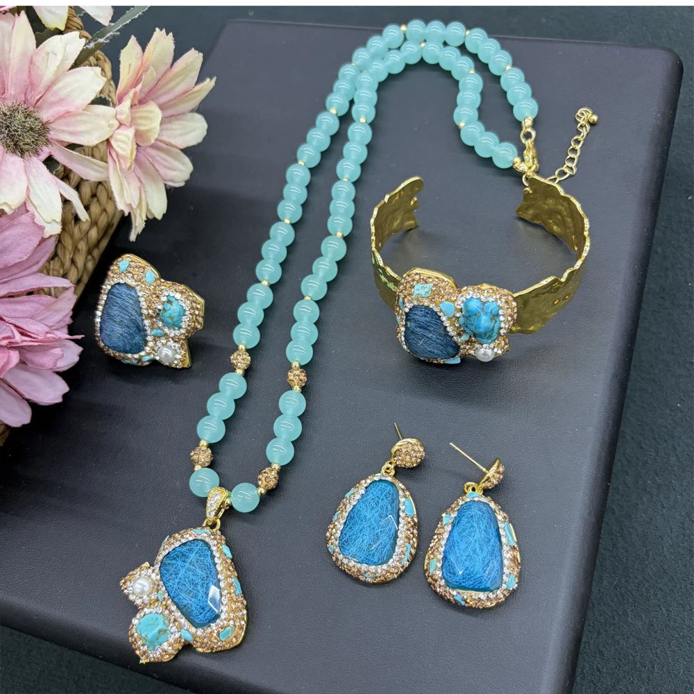A gold jewelry set featuring a beaded necklace, cuff bracelet, ring, and drop earrings with blue stone inlays, displayed on a dark gray surface with decorative flowers.