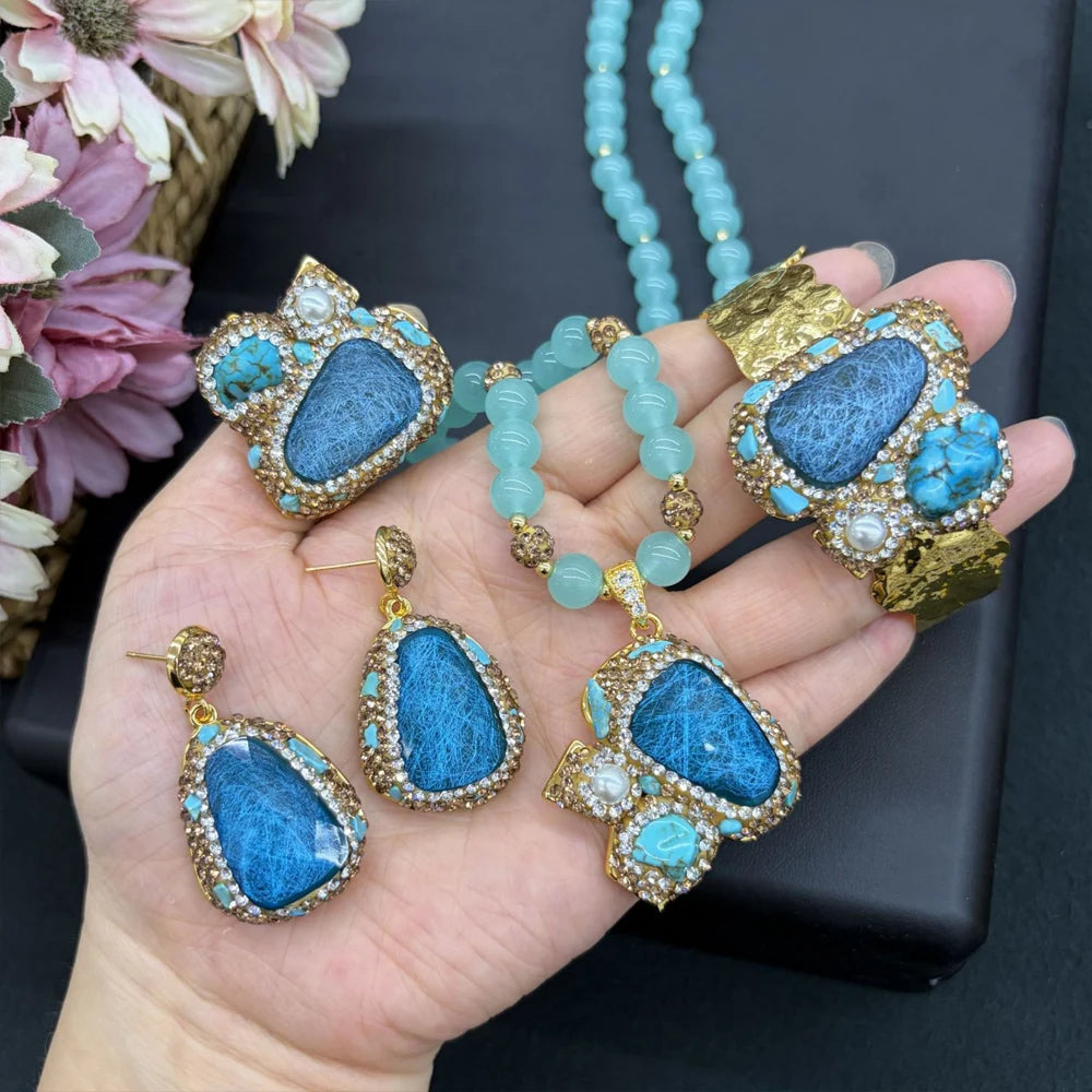 A hand holds a matching jewelry set featuring a turquoise-blue beaded necklace, a large cuff bracelet, a ring, and two teardrop earrings, all decorated with textured blue stones and gold-toned settings with small white stones.