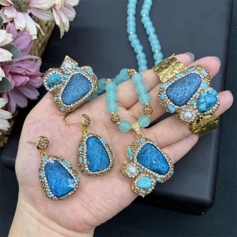 A hand holds a matching jewelry set featuring a turquoise-blue beaded necklace, a large cuff bracelet, a ring, and two teardrop earrings, all decorated with textured blue stones and gold-toned settings with small white stones.