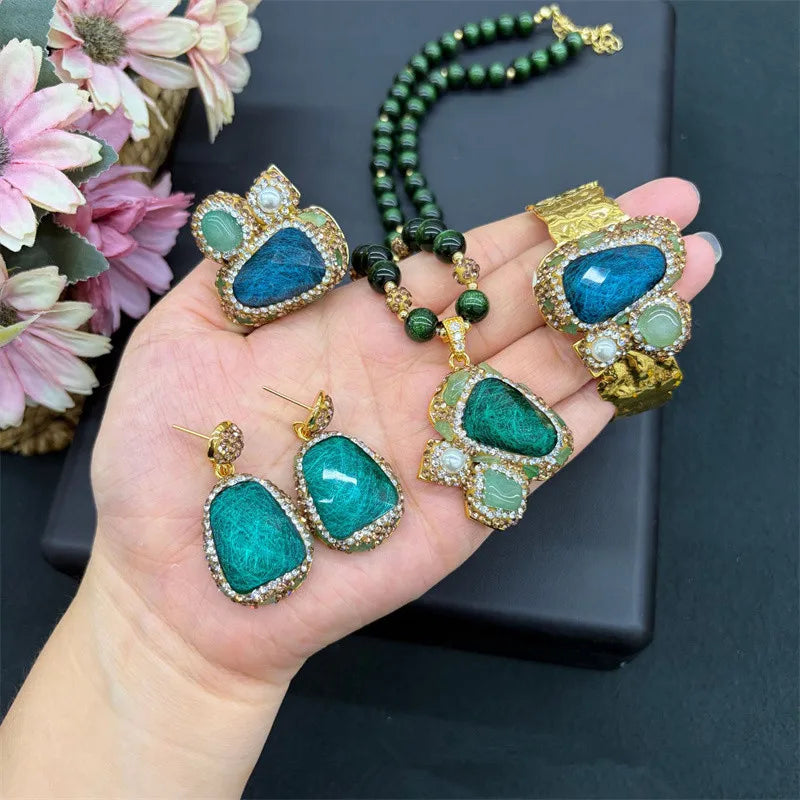 A jewelry set displayed on a hand, featuring a green beaded necklace, a large gold and turquoise necklace, matching earrings, and a gold ring, with pink flowers in the background.
