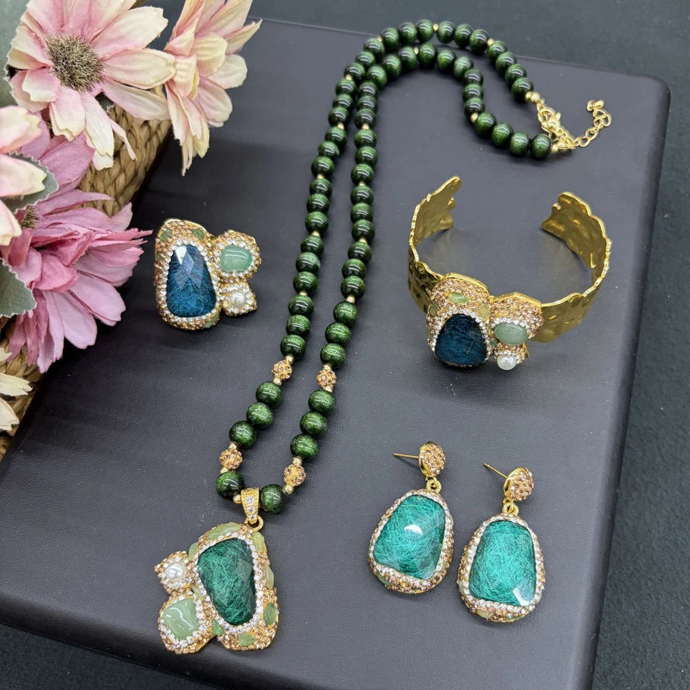 A jewelry set displayed on a dark gray surface, featuring a long beaded necklace, a cuff bracelet, and two matching drop earrings. The pieces combine deep green stones with gold-toned settings and small light green stones.