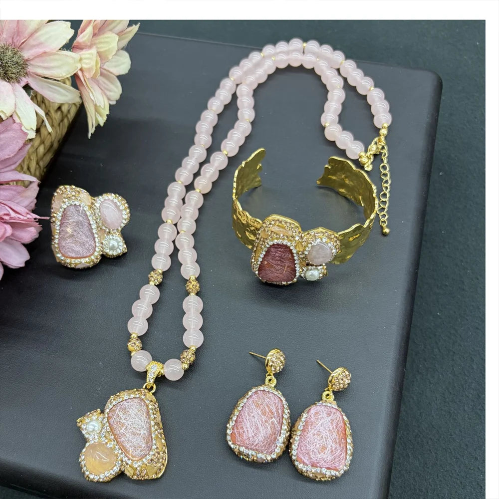 A jewelry set displayed on a dark gray surface, featuring a gold-tone cuff bracelet, matching pendant necklace, and two matching drop earrings. The jewelry is decorated with light pink beads, gold-tone filigree, and pink stone inlays.